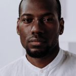 Home Headshot of a serious black man in a white shirt, indoors with a neutral background.